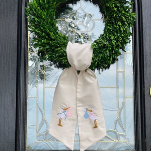 May include: A white fabric door wreath with two embroidered storks carrying a pink and blue bundle. The wreath is hung on a black door with a glass window.