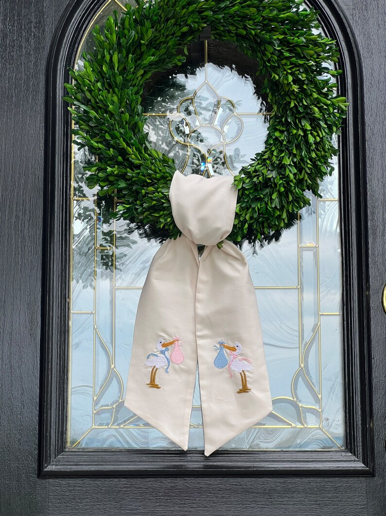 May include: A white fabric door wreath with two embroidered storks carrying a pink and blue bundle. The wreath is hung on a black door with a glass window.