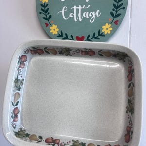 May include: A square, off-white speckled ceramic baking dish featuring a decorative border of colourful pears, apples, and green foliage along the inner rim. The dish has a smooth, glossy finish and rounded corners, designed for oven use.