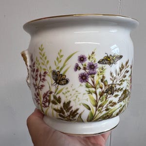 Puede incluir: Una maceta de cerámica blanca con una ilustración botánica de flores silvestres moradas, hierbas verdes y varias polillas y mariposas. El recipiente tiene un cuerpo redondeado, un borde acampanado y un borde dorado en la parte superior.