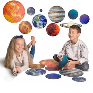 Könnte beinhalten: Zwei Kinder spielen mit einem Satz runder, bunter Planeten-Bodenmatten aus Filz. Die Matten zeigen Bilder von Sonne, Erde, Mars und Jupiter. Die Kinder lächeln und haben Spaß.