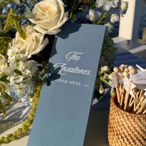 May include: A tall, rectangular lake blue linen drink menu card with white text reading The Anzalones DRINK MENU, positioned next to a woven basket filled with wooden drink stirrers featuring small white flags with the same name printed on them.