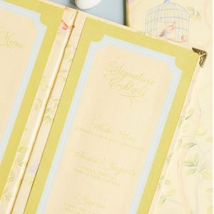 May include: A tri-fold menu card featuring a pale yellow background with a light green border and a delicate birdcage and bird illustration. The text reads Menu, Signature Cocktails, Whiskey Sour, Playful Margarita, and Meertini with ingredients.