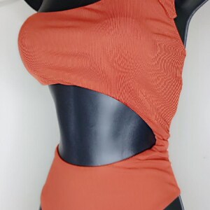 May include: A burnt orange one-piece swimsuit with a one-shoulder design and a side cutout. The swimsuit has a ribbed texture and a tie detail on the shoulder. The swimsuit is displayed on a black mannequin.