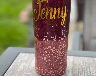 May include: A personalized tumbler with a glittery burgundy and rose gold design. The name "Jenny" is displayed in gold lettering. The tumbler has a clear lid and a straw.