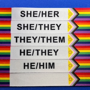 May include: Five white ribbons with black text indicating pronouns: SHE/HER, SHE/THEY, THEY/THEM, HE/THEY, and HE/HIM. Each ribbon has a rainbow and a transgender flag design on the right side.