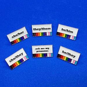 May include: A selection of enamel pins with white rectangular designs and silver edges. Each pin features a pronoun set: "she/her", "they/them", "he/him", "she/they", and "he/they". One pin reads "ask me my pronouns". Each pin has a rainbow stripe design.