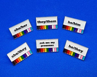 May include: A collection of enamel pins with white rectangular backgrounds and silver borders. Each pin displays a pronoun set: "she/her", "they/them", "he/him", "she/they", and "he/they". One pin reads "ask me my pronouns". Each pin features a rainbow stripe design.