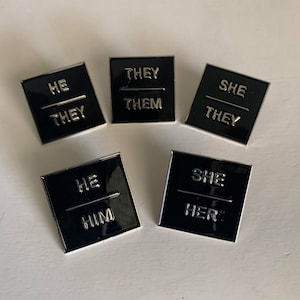 May include: Five black enamel pins with silver lettering. Each pin features a different pronoun set: He/They, They/Them, She/They, He/Him, She/Her.