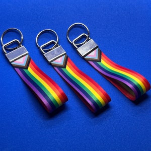May include: Three keyrings with rainbow-striped straps, each with a silver metal clasp and a Pride flag design. The straps show the colours of the rainbow, with a section of the Progress Pride flag. The keyrings are set against a blue background.