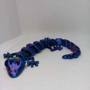 May include: A 3D-printed articulated lizard toy in shades of blue and purple. The toy is designed with multiple segments, allowing for flexible movement. The lizard is posed on a white surface.