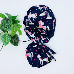 May include: A black scrub hat with a tie, featuring a colorful unicorn print on a dark blue background. The hat has a white star pattern and pink, blue, and yellow accents.