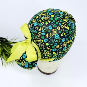 May include: A patterned scrub cap with a bright yellow bow. The cap has a black background with a repeating pattern of yellow, green, and teal shapes. The bow is a solid, vibrant yellow. The cap is displayed on a mannequin head.