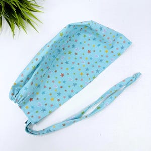 May include: A light blue surgical scrub cap with a pattern of colorful stars and hearts. The cap has a tie in the back for an adjustable fit. The fabric is a soft cotton material.