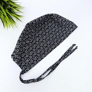 May include: A black and white patterned surgical scrub cap with a tie closure. The cap features a repeating pattern of white circles on a black background. The ties are made of the same fabric.