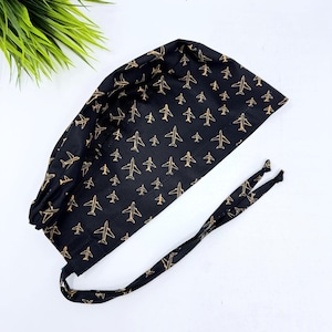 May include: Black surgical scrub cap with a repeating pattern of gold-colored airplanes. The cap has a tie closure at the back and is made of a soft, flexible fabric. The cap is designed to cover the hair during medical procedures.