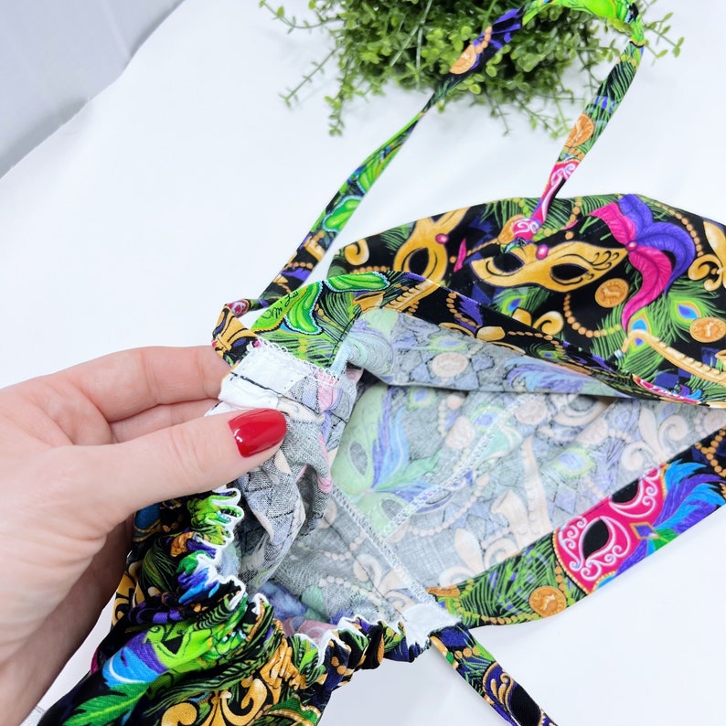 May include: A colorful fabric bag with a Mardi Gras mask pattern. The bag features a black background with gold, green, purple, and pink mask designs. The interior lining has a blue, green, and white pattern. The bag has a drawstring closure and straps.