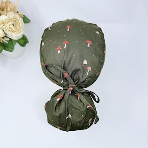 May include: A green scrub hat with a white and red mushroom print. The hat has a tie at the back.