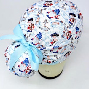 May include: A white scrub cap with a nautical gnome pattern, featuring gnomes, anchors, and ships' wheels in red, blue, and white. A light blue ribbon is tied in a bow on the side. The cap is designed for medical professionals.
