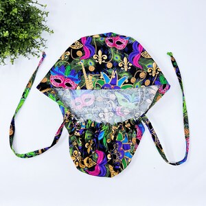May include: A black surgical scrub cap with a vibrant pattern of Mardi Gras masks, fleur-de-lis, and peacock feathers in purple, green, blue, and gold. The cap has ties for a secure fit and is made of a soft fabric.
