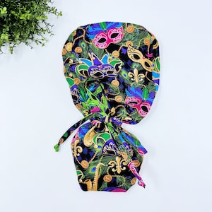 May include: A black scrub cap with a colorful Mardi Gras mask pattern. The cap features masks in pink, gold, purple, and green, along with gold coins and fleur-de-lis symbols. The cap has a drawstring closure.