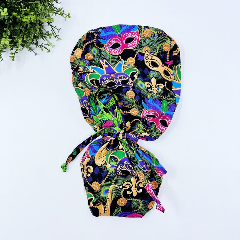 May include: A black scrub cap with a colorful Mardi Gras mask pattern. The cap features masks in pink, gold, purple, and green, along with gold coins and fleur-de-lis symbols. The cap has a drawstring closure.