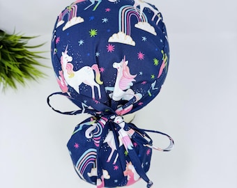 May include: A navy blue scrub cap featuring a whimsical unicorn and rainbow pattern. The cap has a ponytail style with a matching fabric tie. The design includes white unicorns, rainbows, clouds, and stars in pink, yellow, and blue.
