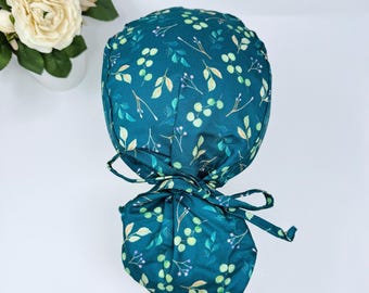 May include: A teal scrub cap with a botanical pattern of green leaves and berries. The cap is tied at the back with matching fabric ties. The design features a dark teal background with light green and yellow accents.