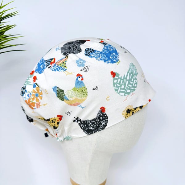 May include: White fabric cap featuring a colorful chicken pattern. The chickens are depicted in various colors and patterns, including blue, green, yellow, and black. The cap is designed to fit the head and has a rounded shape.