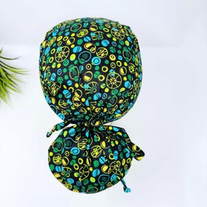 Op de afbeelding: Een zwarte scrubcap met een kleurrijk patroon van gele, groene en blauwe vormen. De cap is aan de achterkant vastgebonden met een bijpassende stoffen stropdas. Het ontwerp omvat cirkels, ovalen en wielachtige patronen.