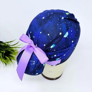 May include: A blue scrub cap with a space-themed pattern featuring white stars and planets. The cap is adorned with a lavender bow. This cap is designed for medical professionals and is made of a soft, comfortable material.
