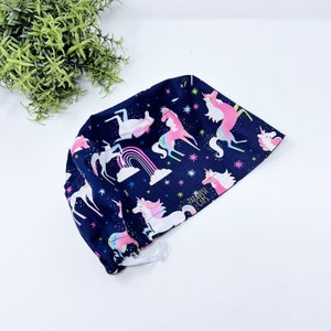 May include: A navy blue surgical scrub cap featuring a whimsical pattern of unicorns, rainbows, and stars. The unicorns are depicted in shades of pink, white, and blue. The text "Paradise Caps" is printed on the side. The cap has an elastic band.
