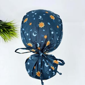 May include: A dark blue scrub cap featuring a celestial design of yellow suns, white moons, and stars. The cap has a tie closure at the back. It is designed for use in medical or healthcare environments.