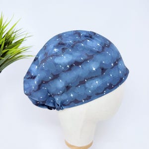May include: A blue scrub cap featuring a repeating pattern of dark blue clouds and small white four-pointed stars. The fabric head covering has a rounded crown and a fitted band, designed to be worn securely around the head for medical or work use.