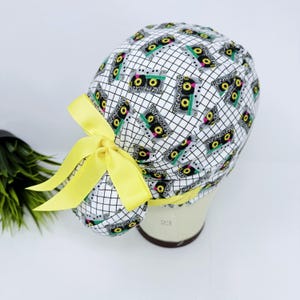 May include: A white scrub cap featuring a black grid pattern and a repeating design of cassette tapes in black, green, yellow, and pink. The cap is tied with a bright yellow bow.