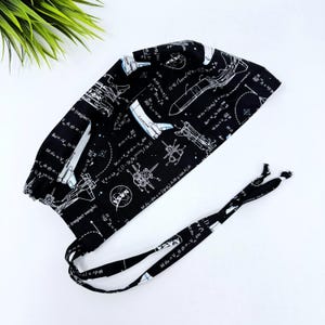 May include: Black surgical scrub cap with a space-themed pattern. The cap features white illustrations of rockets, space shuttles, and mathematical equations on a black background. The NASA logo is also visible. The cap has ties for a secure fit.