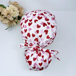 May include: A white scrub cap with a pattern of red and pink hearts. The cap is tied at the back with a matching bow. The fabric is soft and the design is perfect for Valentine's Day or any occasion.