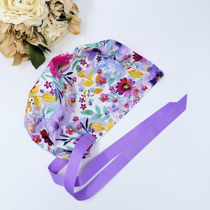 May include: A scrub cap with a floral pattern in shades of purple, pink, yellow, and blue on a white base. It has a lavender ribbon tie. The cap is designed for medical professionals.