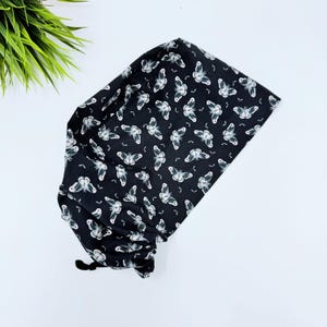 May include: Black scrub hat with a white moth pattern. The hat has a drawstring tie.