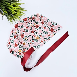 Puede incluir: Un gorro de quirófano blanco con estampado floral en tonos rosa, rojo y verde. El gorro tiene dos cintas largas de color burdeos.