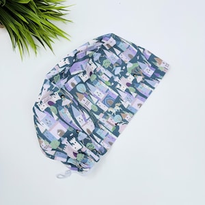 Puede incluir: Un gorro de quirófano azul con un cordón blanco. El gorro tiene un patrón repetido de castillos rosas y morados, árboles y personas.