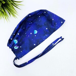 May include: A navy blue surgical scrub cap with a space-themed pattern. The cap features white stars and small, colorful spaceship illustrations. It has long ties for a secure fit and is made of a soft, flexible fabric.