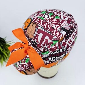 May include: A white and maroon patterned scrub cap with orange bow. The cap features the text "Texas A&M Aggies" and other related graphics. The bow is made of a wide, solid orange ribbon.