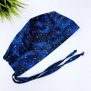 May include: A dark blue surgical scrub cap with a celestial constellation pattern. The cap features white star constellations against a dark blue background, with matching ties for secure fastening. The cap is designed for medical professionals.