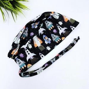 May include: Black surgical scrub cap with a colorful space-themed pattern. The cap features rockets, planets, and alien figures in shades of white, orange, blue, and purple. The cap has a tie in the back for an adjustable fit.