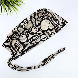 May include: A black and beige skull and skeleton print surgical scrub cap. The cap is made of fabric and has a tie in the back. The pattern includes skulls, rib cages, and other bone structures. The cap is designed for medical professionals.