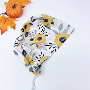 May include: A white fabric scrub hat with a sunflower and floral print. The hat has a drawstring closure.