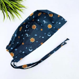 May include: A dark blue scrub cap with a repeating pattern of yellow suns, white moons, and blue stars. The cap has a tie closure and is made of a soft fabric. The cap is designed for medical professionals.