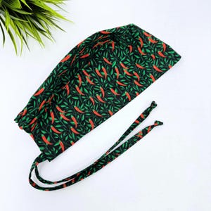 Puede incluir: Un gorro quirúrgico negro con un estampado repetido de chiles rojos y hojas verdes. El gorro tiene un cierre de lazo y está hecho de una tela suave. El fondo es blanco.