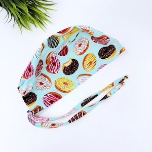 May include: A light blue surgical scrub cap featuring a repeating pattern of colorful donuts with various toppings. The donuts are in shades of brown, yellow, pink, and white. The cap has long ties for securing it.